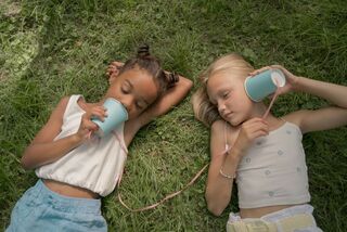 Two young girls lying on grass using homemade cup phones and string to talk to each other outdoors. 