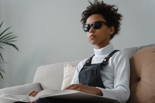 A girl with dark sunglasses is sitting on a couch reading a Braile book.