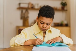 A young boy with curly hair focused on writing in a blue notebook.