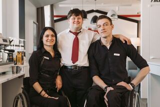 Three workers at a café pose together with smiles. Two of them are in wheelchairs and the third has Down Syndrome.